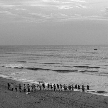 A group of people stands on a beach, looking at the ocean, in a black and white photo.